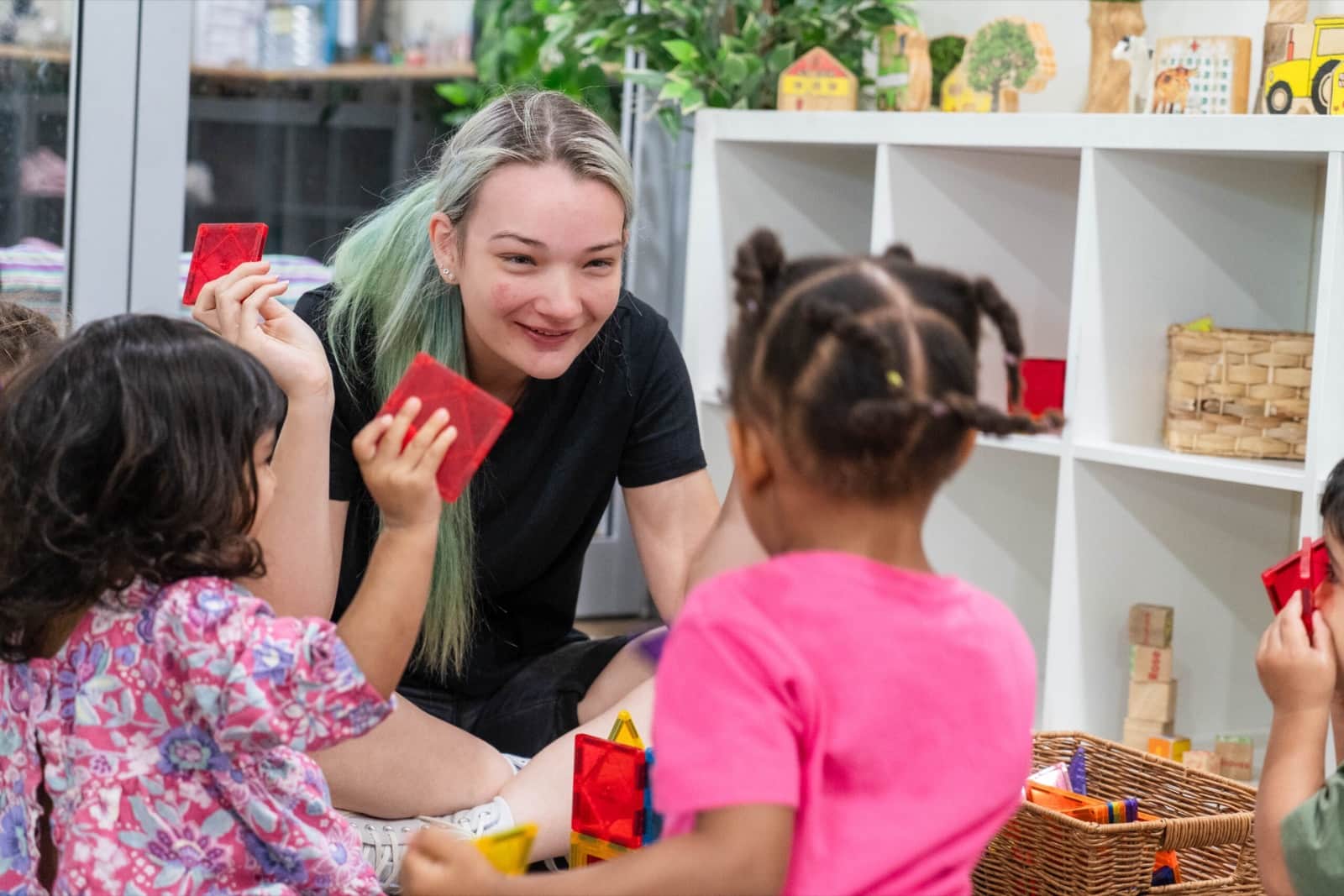 A smiling Saige educator on the floor playing with two little girls and colour blocks